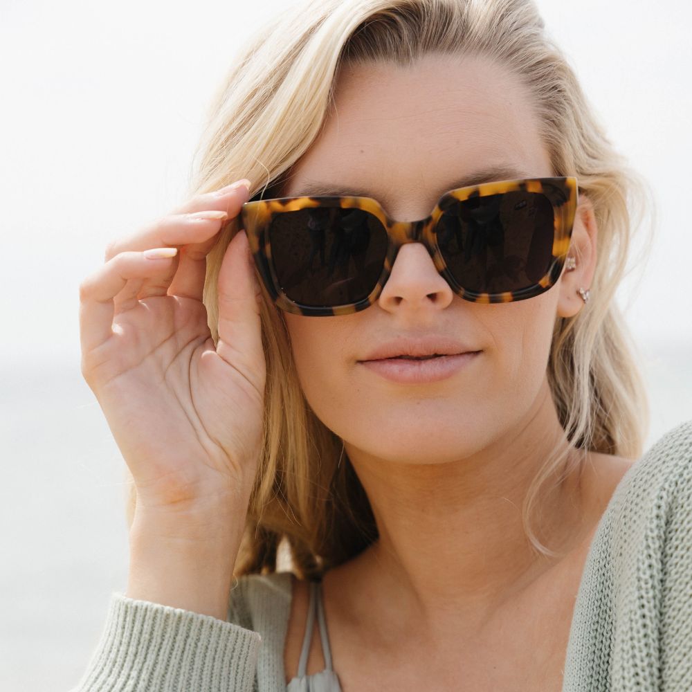 A woman with long blonde hair lifting the Soluna sunglasses, worn outdoors