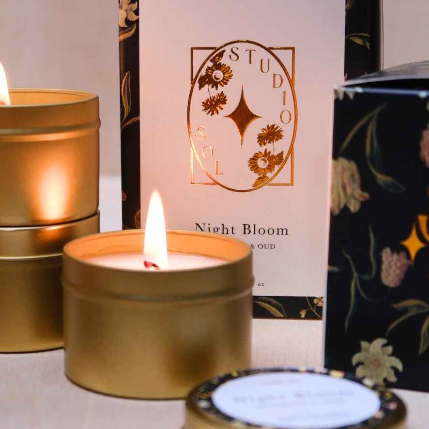 A lit gold travel tin in front of the Night Bloom cartons and a floral panel.