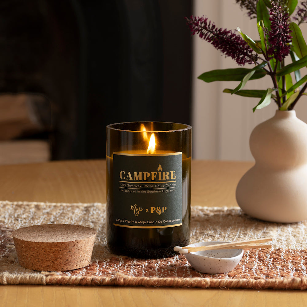 Lit Campfire candle on a woven runner beside a ribbed cream vase of purple blooms, cork lid alongside
