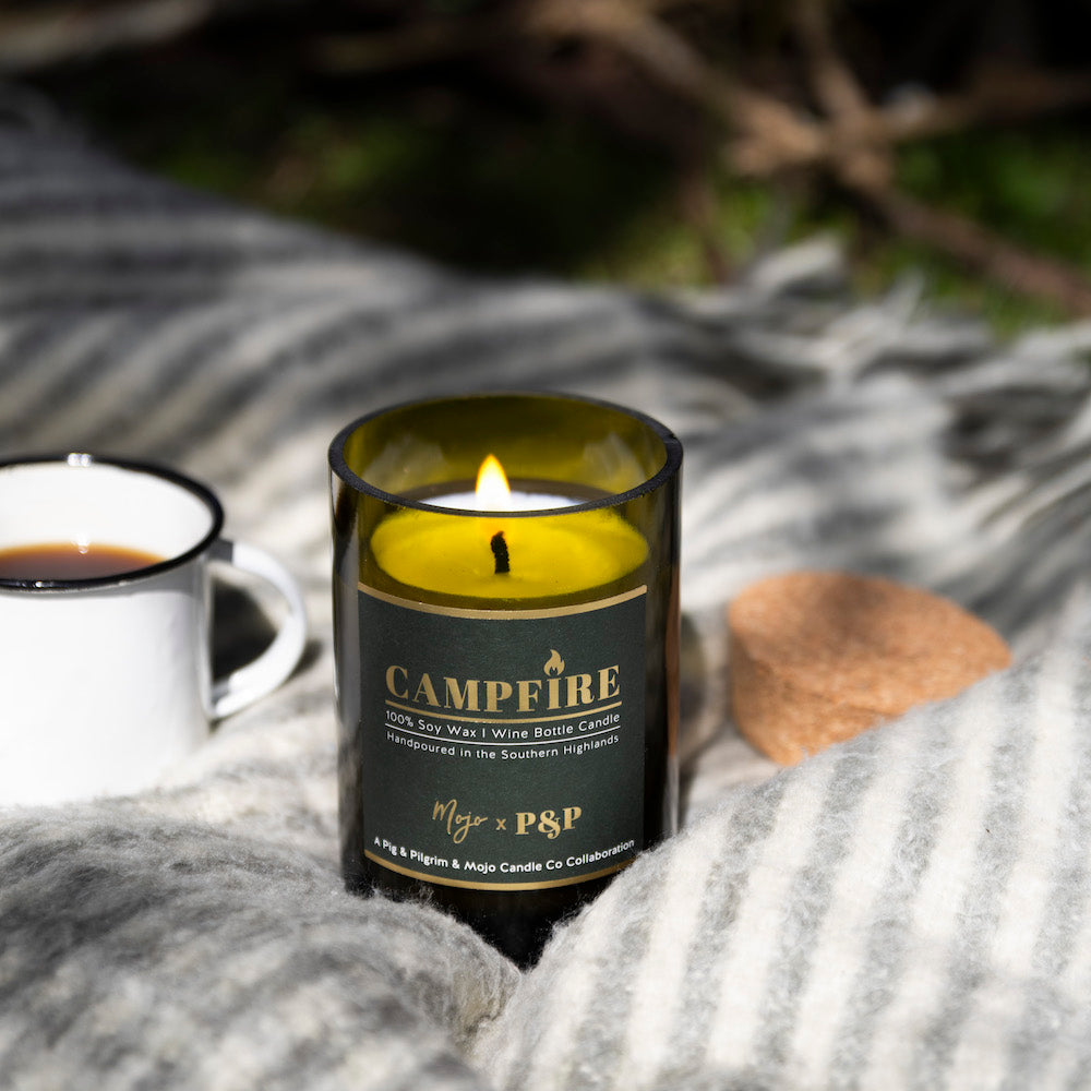 Lit Campfire candle on a striped grey throw with an enamel mug and its cork lid