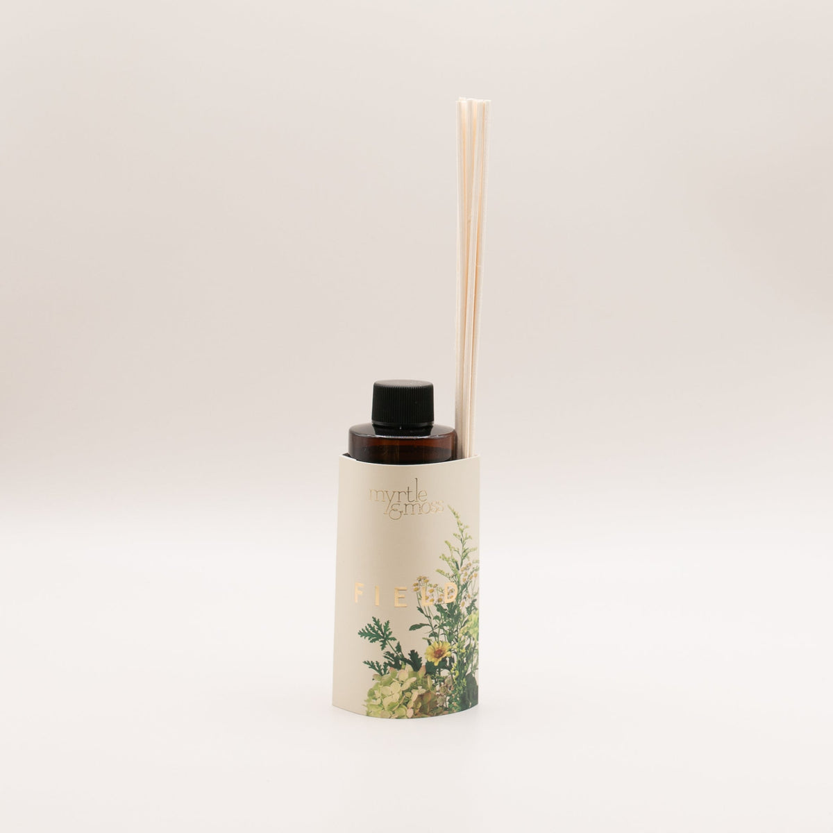 Amber diffuser refill bottle with a black cap in a cream sleeve marked Field in gold, printed with pale green hydrangea, a yellow daisy, small white daisies and green herbs, with rattan reeds tucked into the sleeve, on a pale grey backdrop.