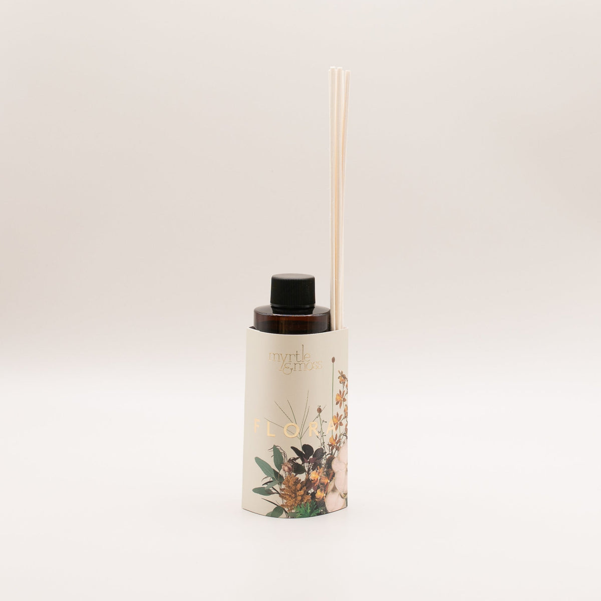 Amber diffuser refill bottle with a black cap in a cream sleeve marked Flora in gold, printed with orange blooms, eucalyptus leaves and a pale blush-white flower, with rattan reeds tucked into the sleeve, on a pale grey backdrop.