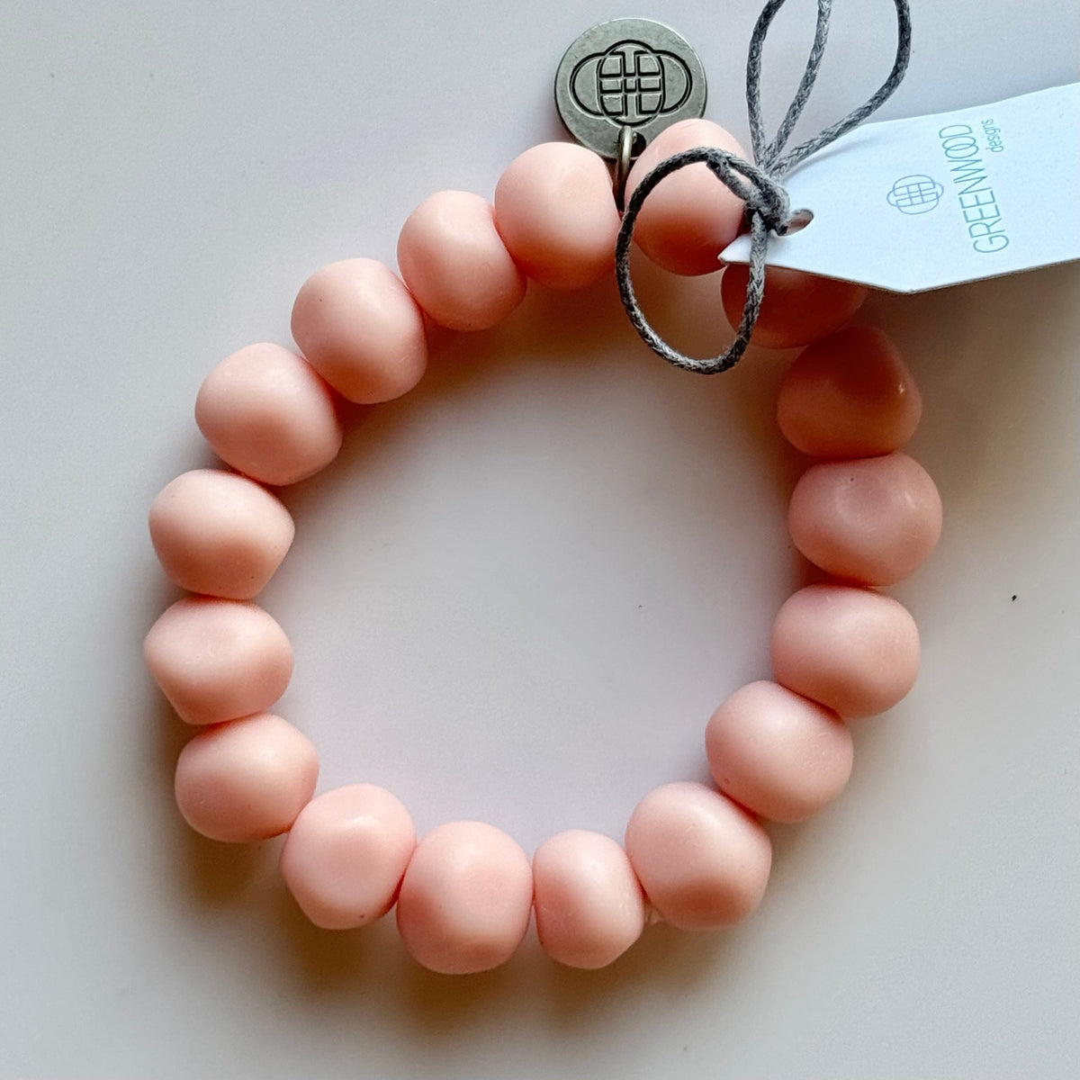 Rock bracelet in Baby Pink: a stretch bracelet of chunky soft peachy-pink resin beads with its swing tag and metal charm, laid on a pale surface.