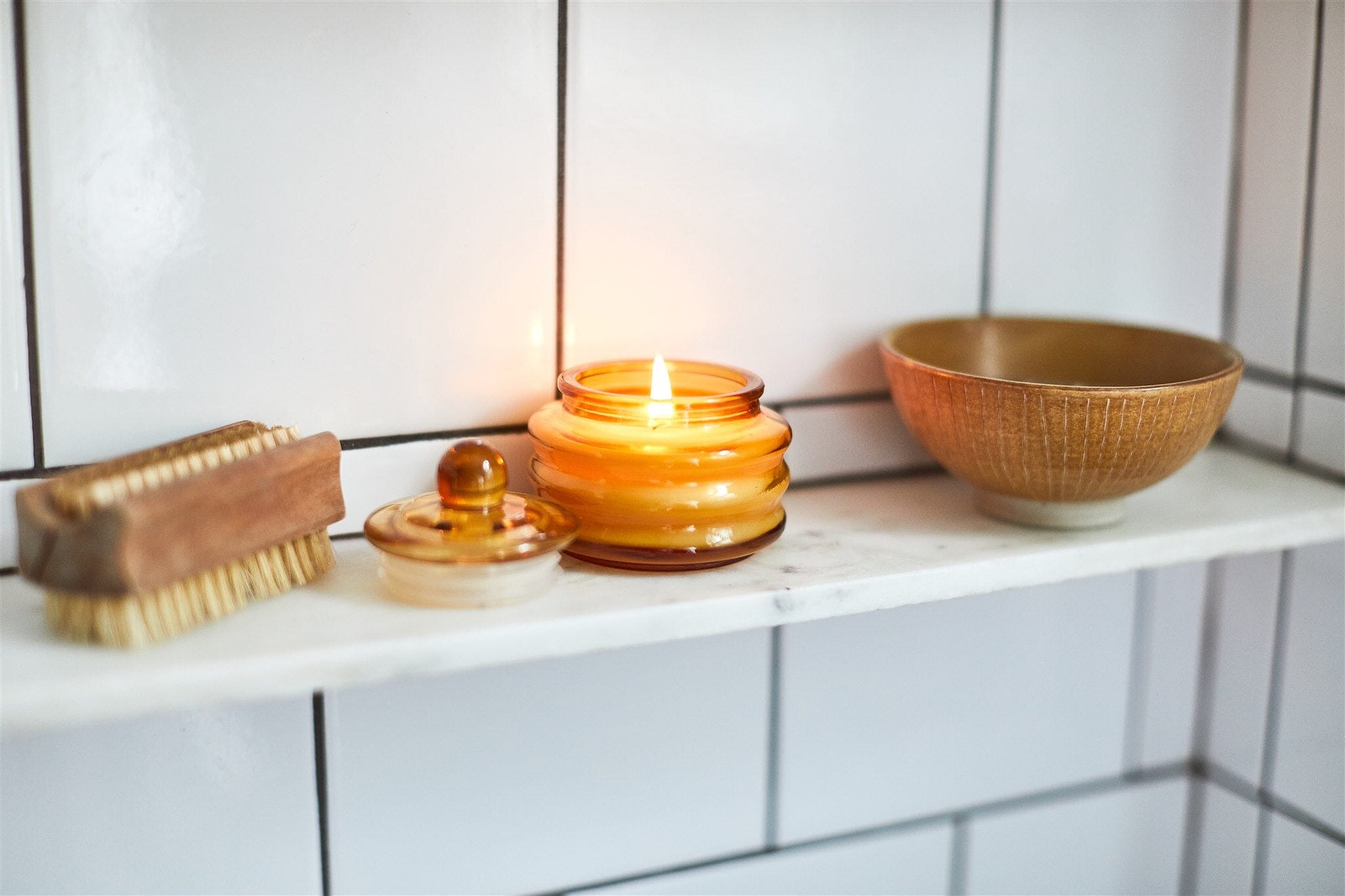 Lit Beam candle glowing amber on a tiled shelf beside a wooden brush and ceramic bowl