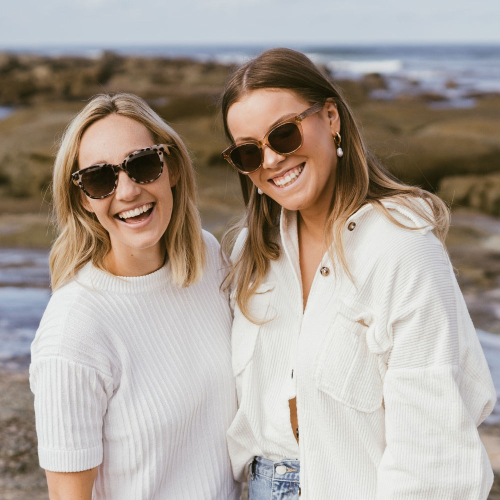 Two women at a rocky beach, both wearing Lila Grace sunglasses