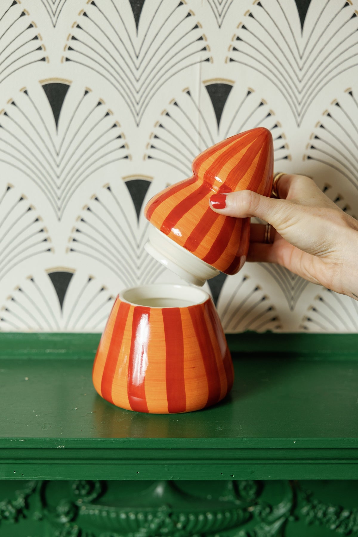 A hand lifting the striped top off the red and orange tree candle on a green mantel, against fan-patterned wallpaper.