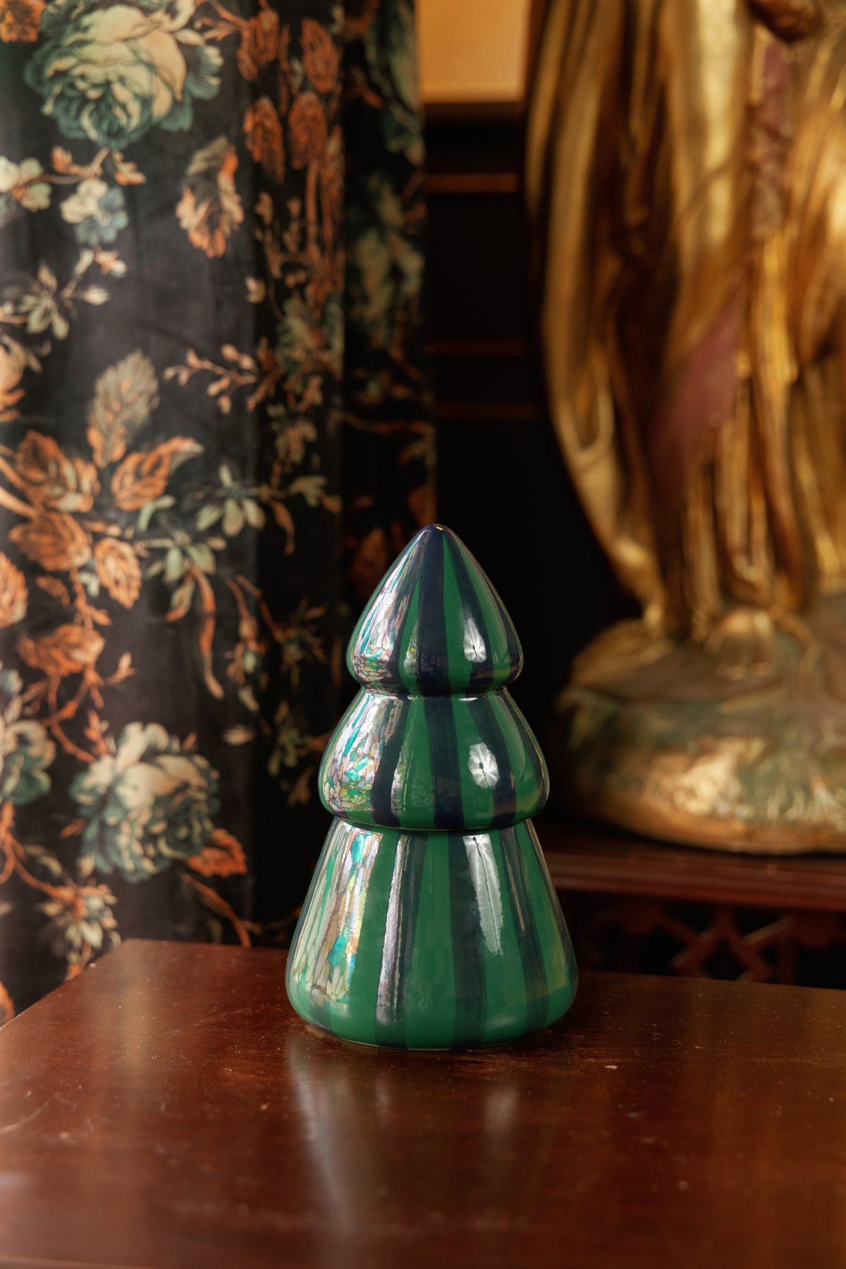 The green and navy striped tree candle on a dark timber side table beside floral curtains and a gilded statue.