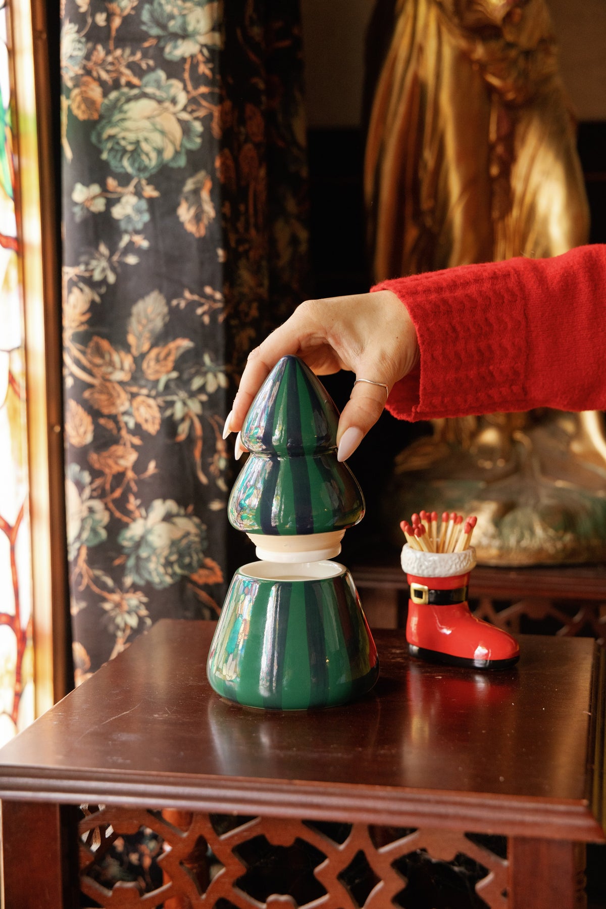 A hand in a red sleeve lifting the top off the green striped tree candle, a red boot of matches beside it.