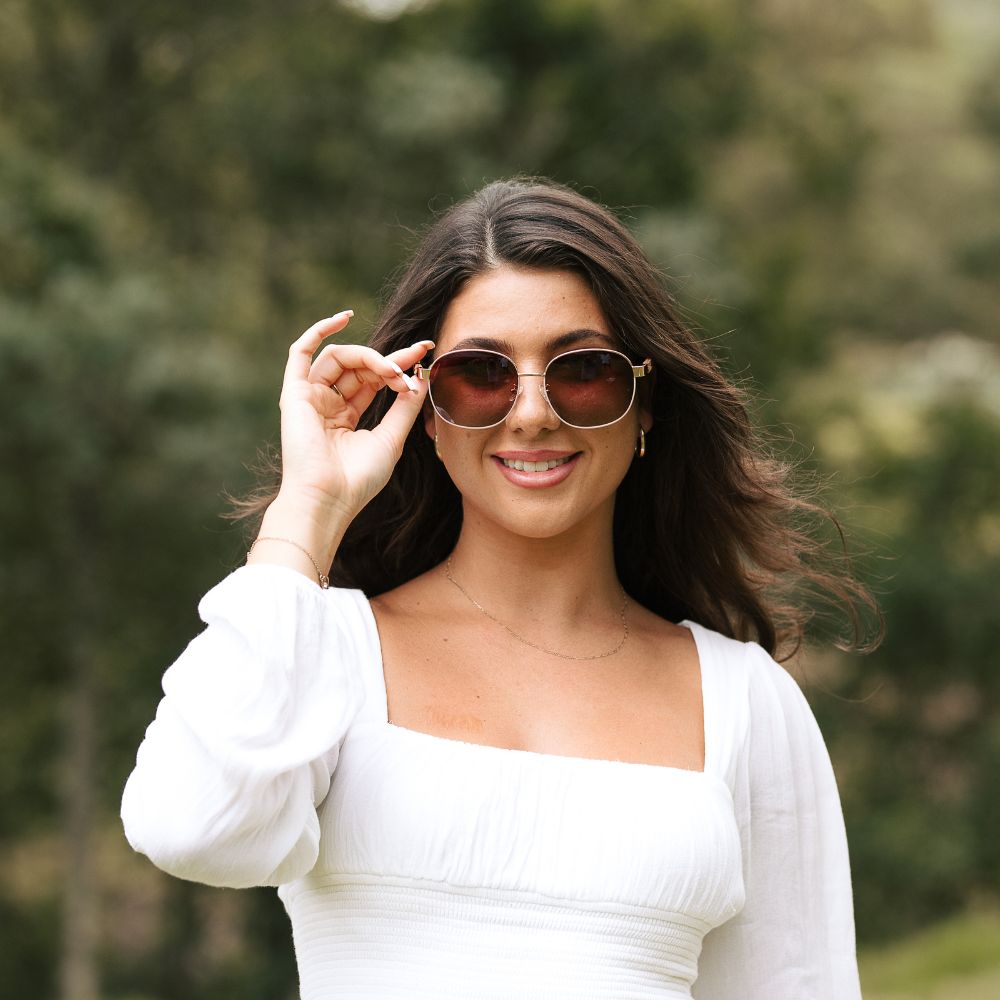A woman in a white top adjusting the Cleo Bloom sunglasses, smiling, with trees behind her