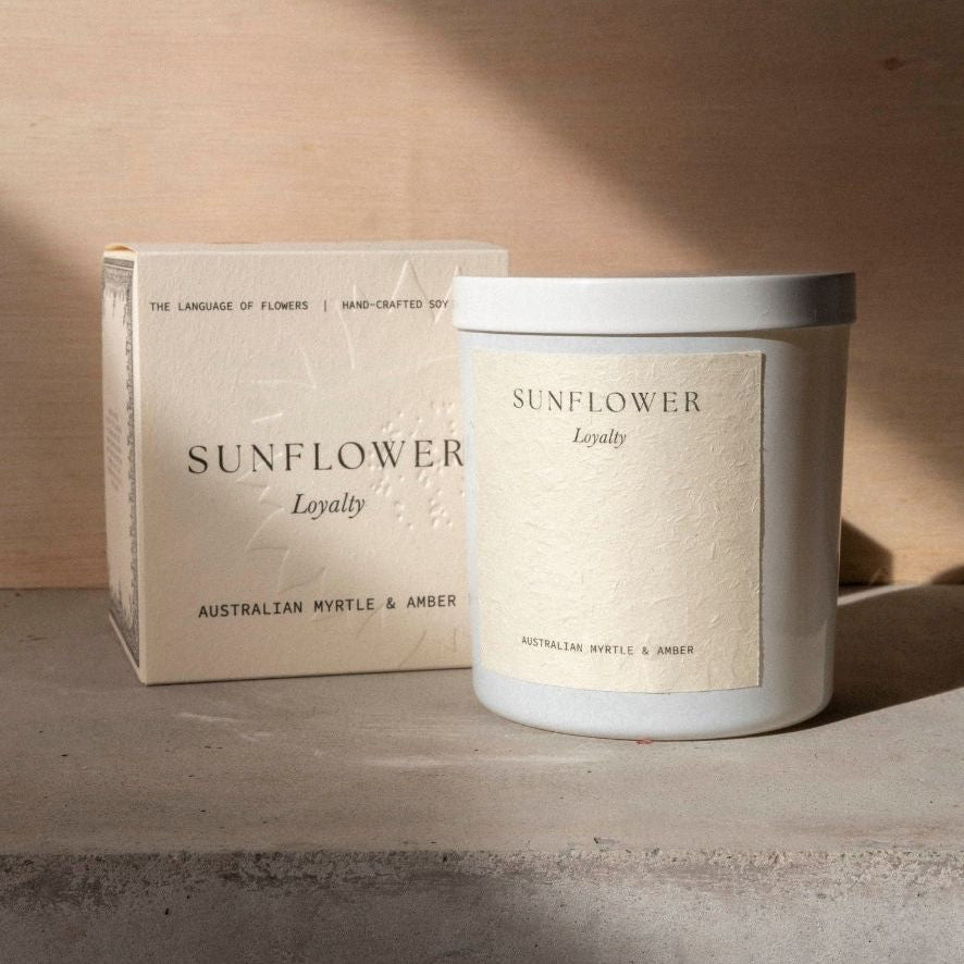 The Sunflower candle in its white vessel beside its cream box on a concrete ledge.