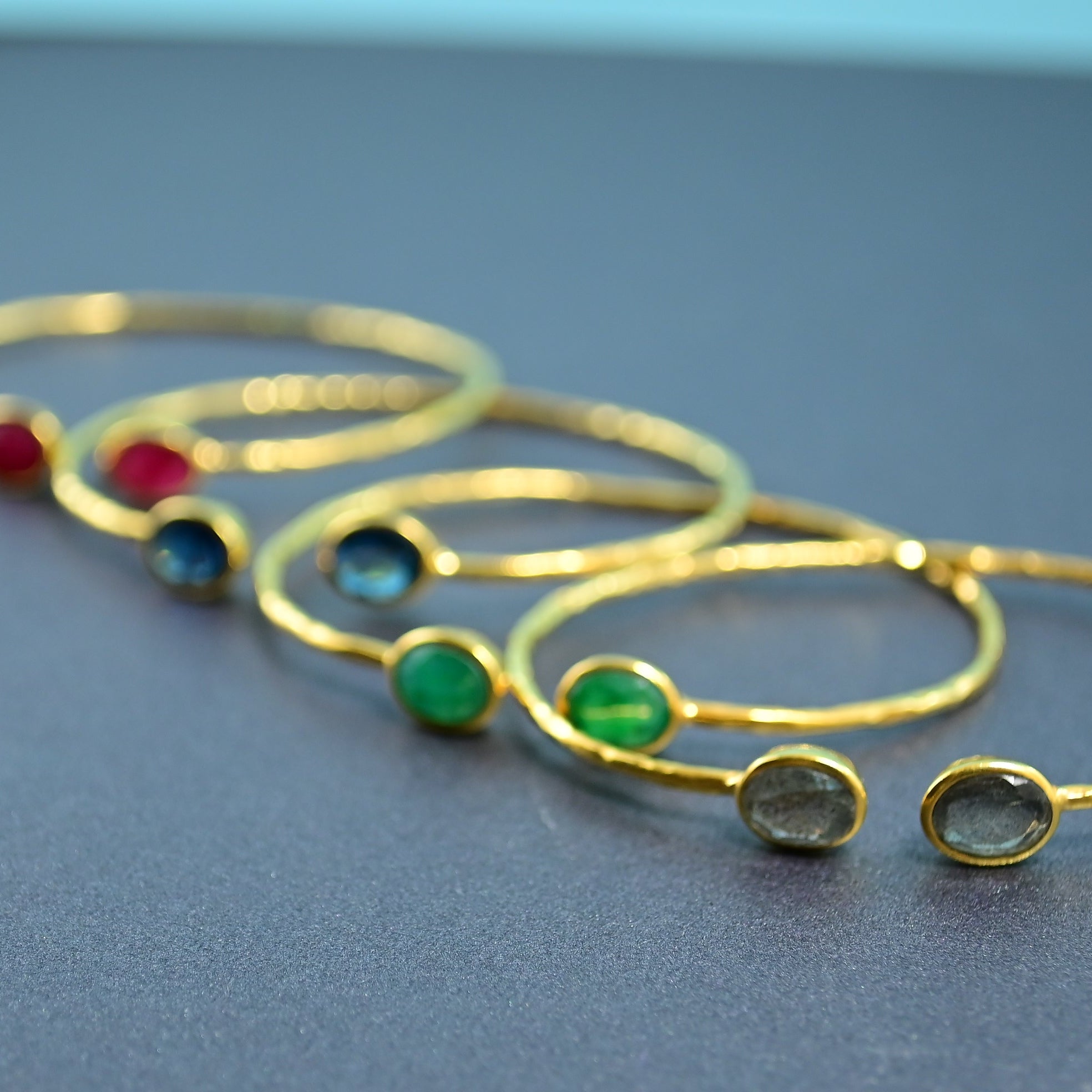 Four Euro Gold gem cuff bangles with ruby, blue topaz, emerald and grey stones on a dark surface