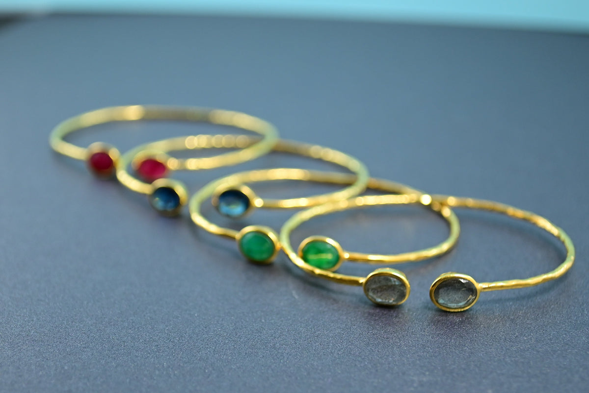 Four Euro Gold gem cuff bangles with ruby, blue topaz, emerald and grey stones on a dark surface