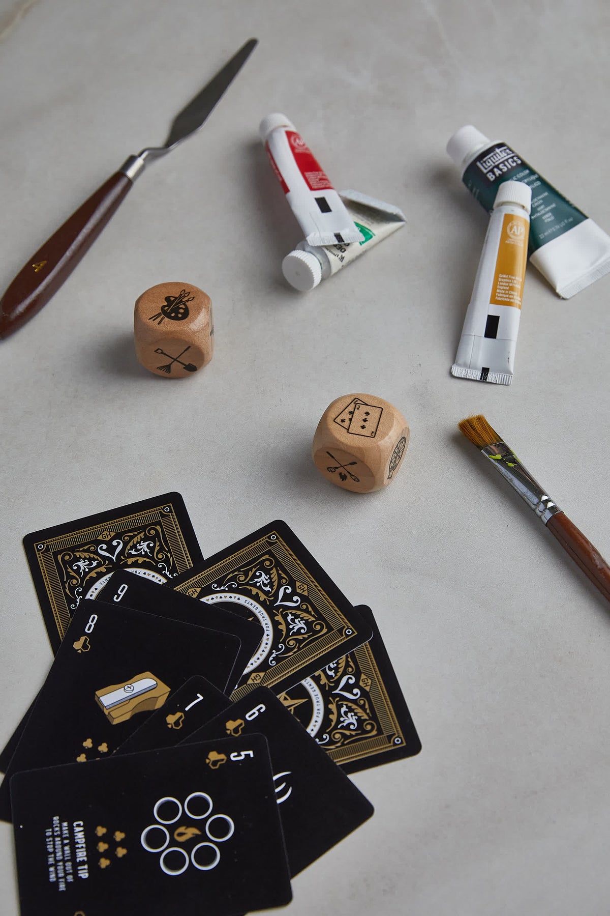 Family Dice with two wooden dice, paint tubes, a palette knife, a paintbrush and playing cards on a light surface.
