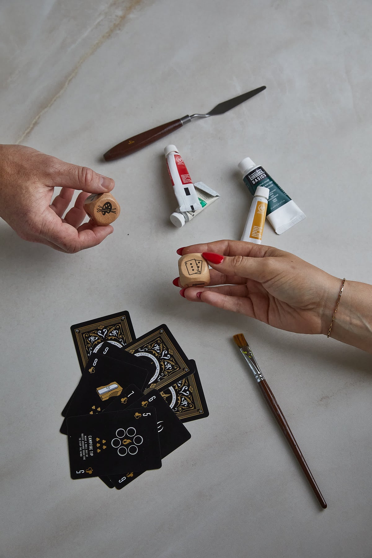 Two hands hold Family Dice over a table with playing cards, paint tubes, a palette knife and a paintbrush.
