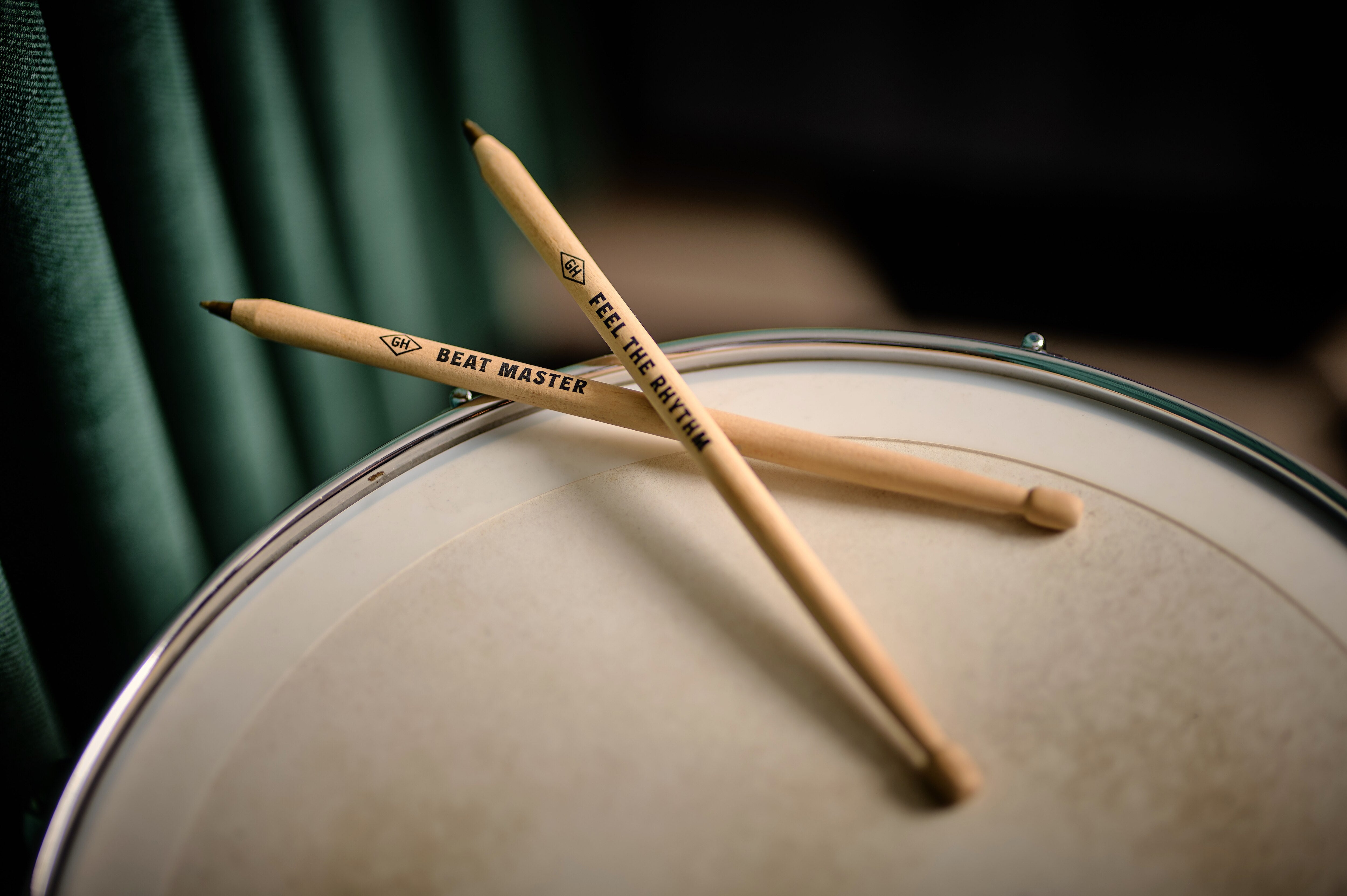 The two drumstick pens resting crossed on a drum skin against a dark green backdrop