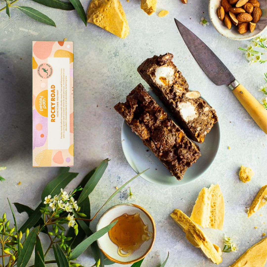 Pebbly Path Honeycomb Highway rocky road 100 g box beside a cut slab on a plate, with honeycomb shards, almonds and honey