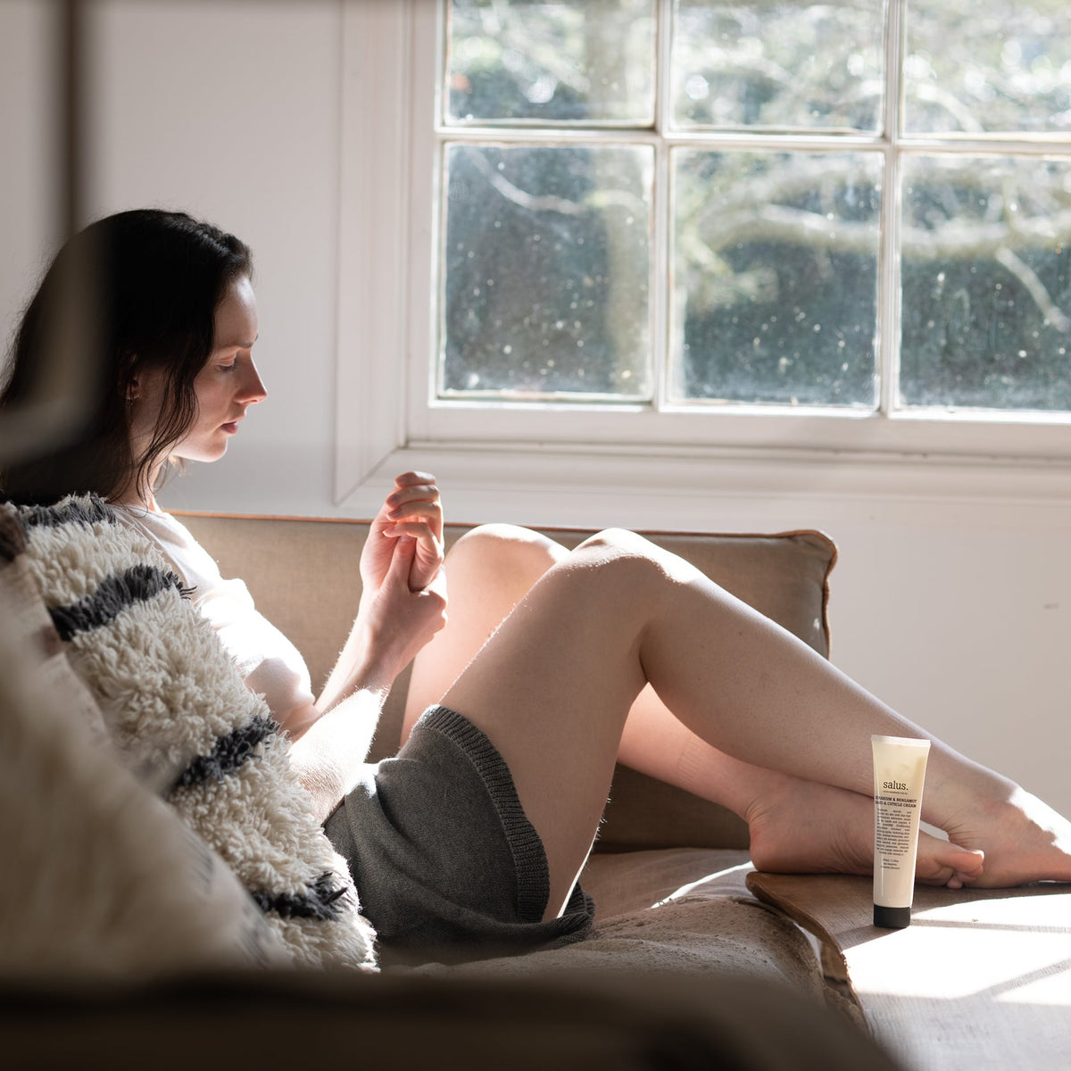 Woman applying Salus Geranium & Bergamot hand cream while relaxing on a sofa in window light