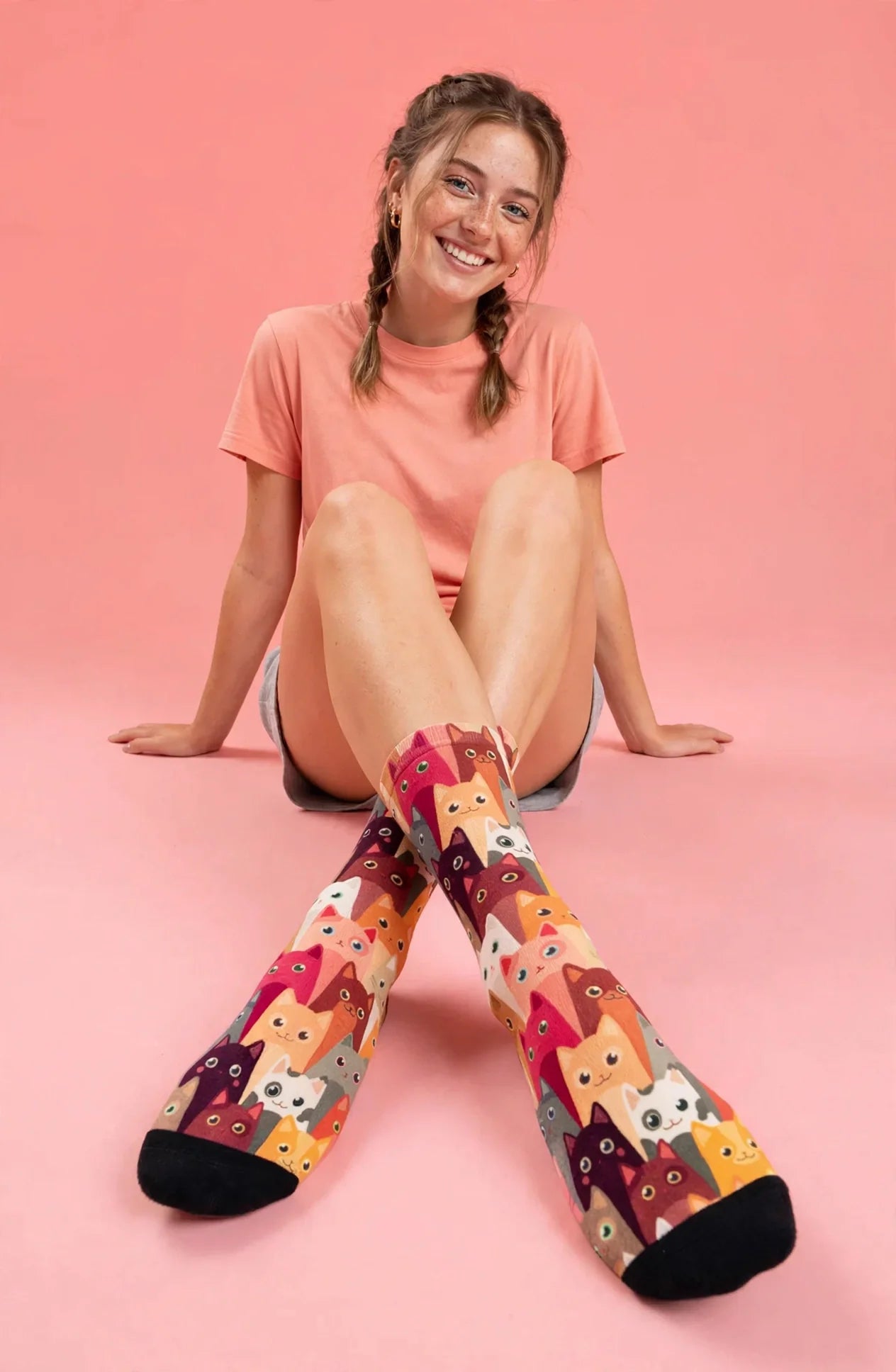Woman in a peach t-shirt sitting on a pink background with her legs crossed, wearing the cat-print socks with black toes.