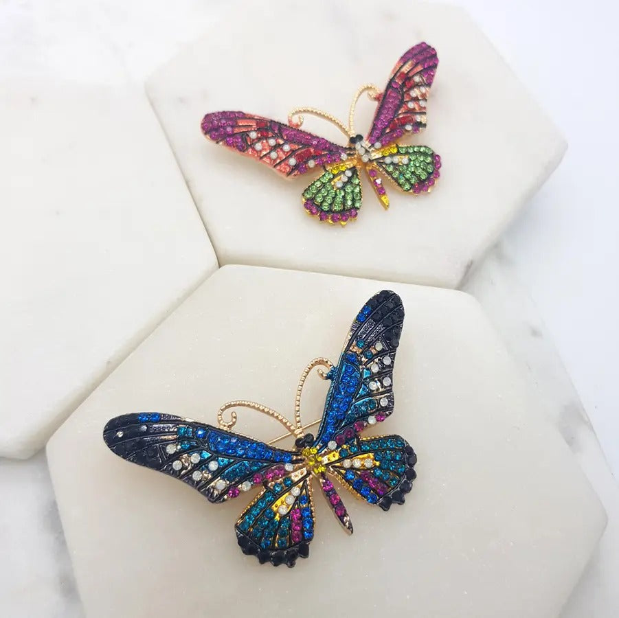 Two crystal butterfly brooches, one in pink and green and one in blue and multicolour, on white marble