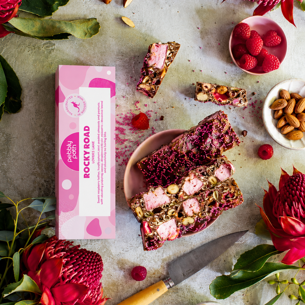 Pebbly Path Lovers Lane rocky road 100 g box with cut slices on a plate, raspberries, almonds and red waratah flowers