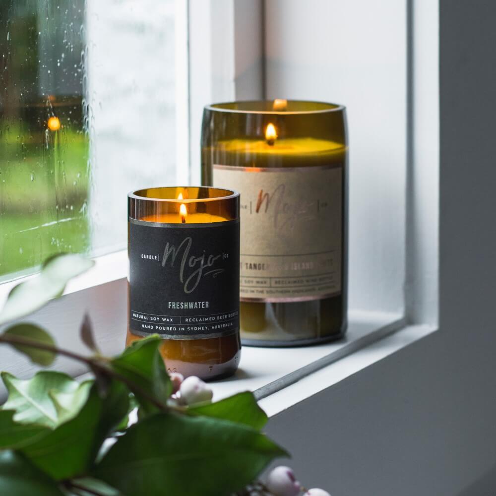 Two lit Mojo bottle candles on a windowsill by a rain-speckled window