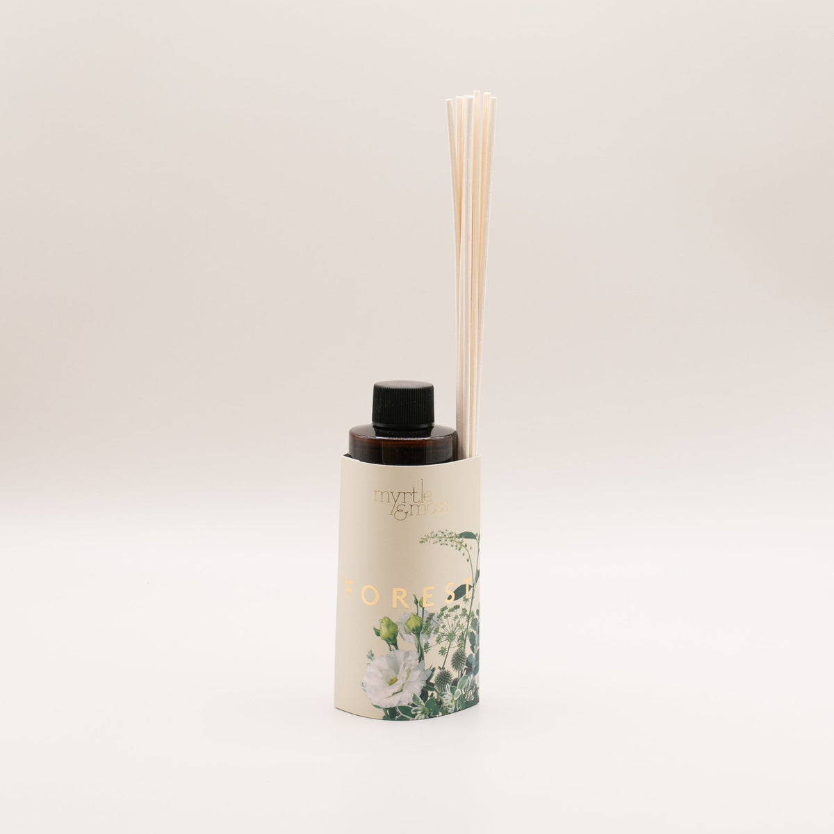Amber diffuser refill bottle with a black cap in a cream sleeve marked Forest in gold, printed with white blooms, green buds and thistles, with rattan reeds tucked into the sleeve, on a pale grey backdrop.