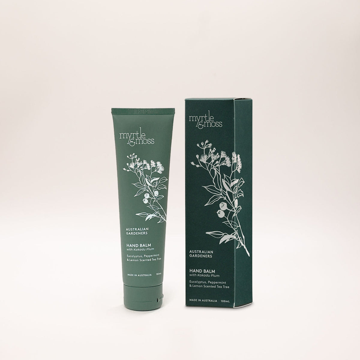 Sage green tube of Australian Gardeners hand balm beside its dark green carton, both printed with a white line drawing of gum blossom, leaves and gumnuts, on a pale backdrop.