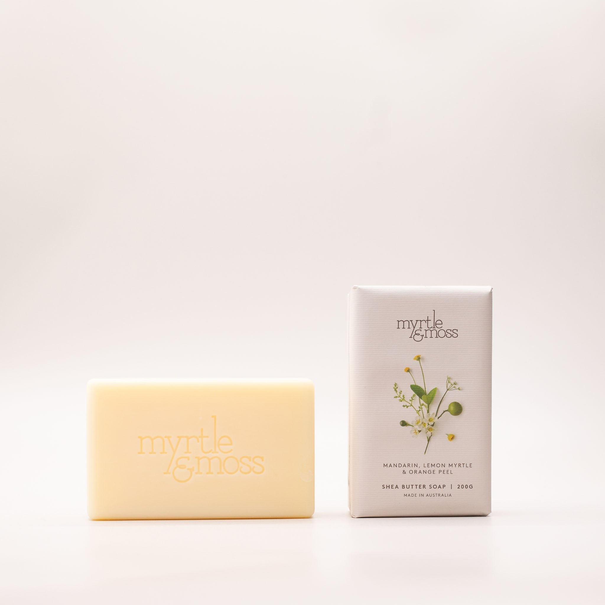 Cream shea butter soap bar embossed 'myrtle & moss' beside its pale grey box, printed with white daisies and yellow buds and labelled Mandarin, Lemon Myrtle & Orange Peel.