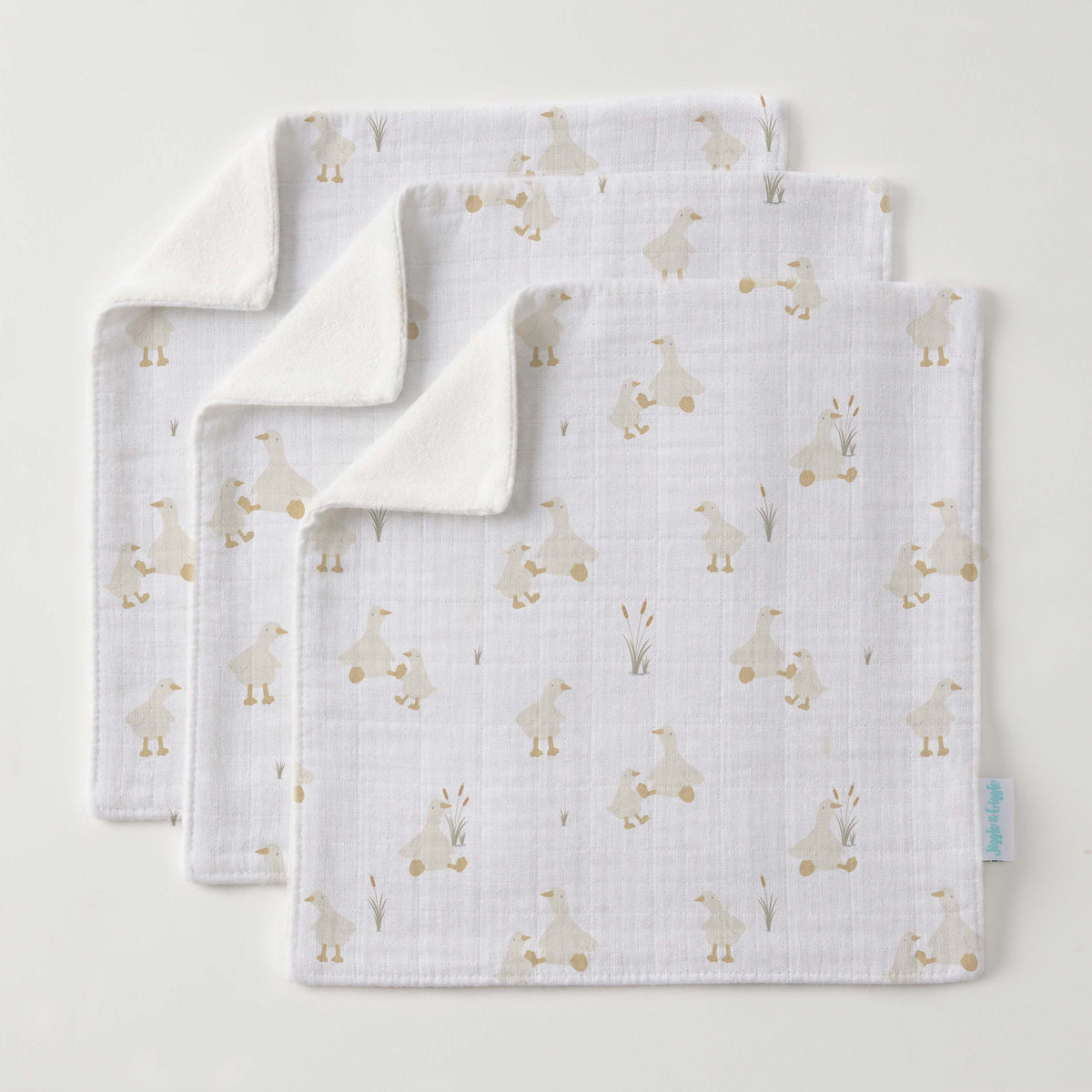 Three white muslin face washers fanned out, printed with small cream ducks and reeds, corners turned back to show soft cream towelling, a small blue tag on one edge