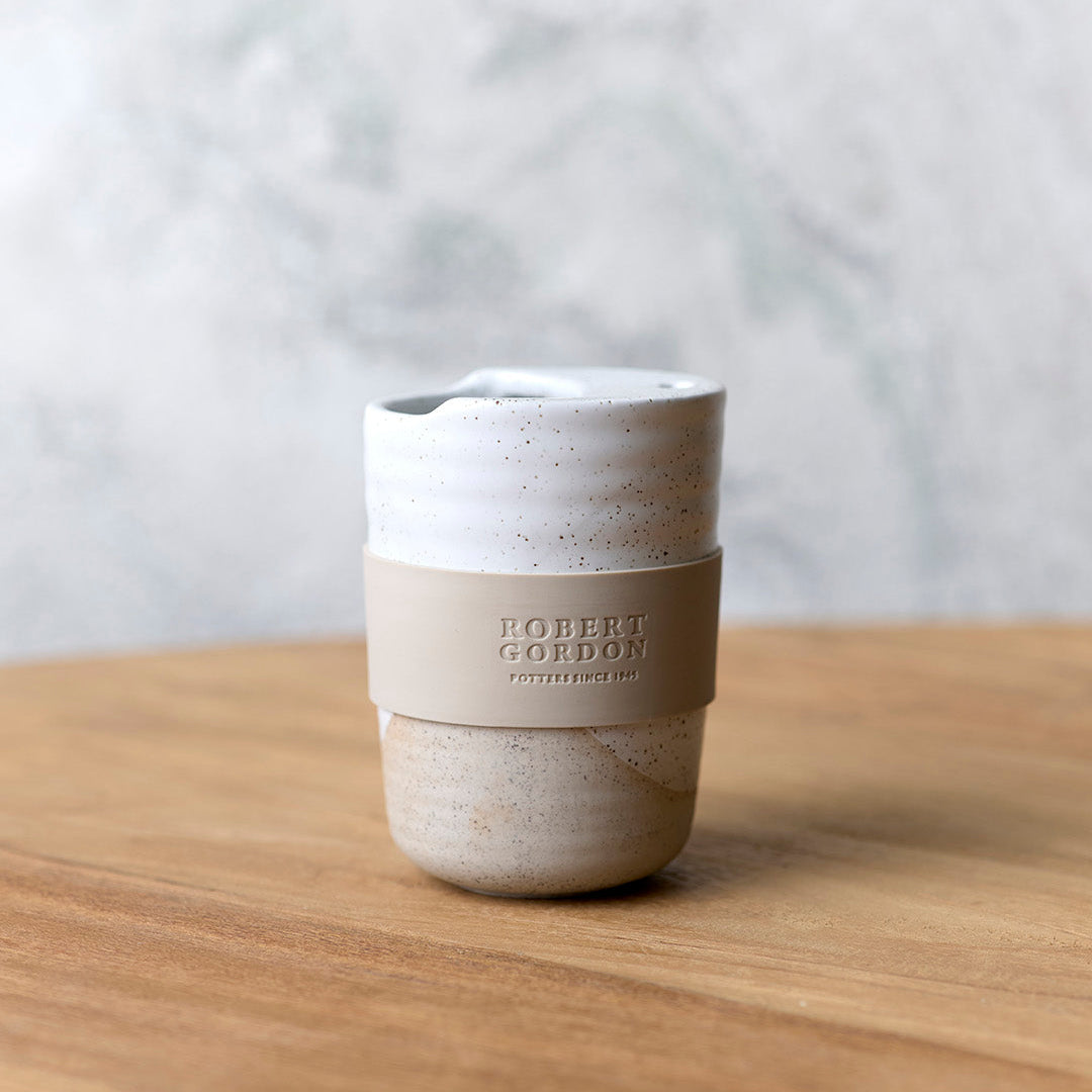White speckle stoneware travel mug with lid and beige silicone band, on a timber bench