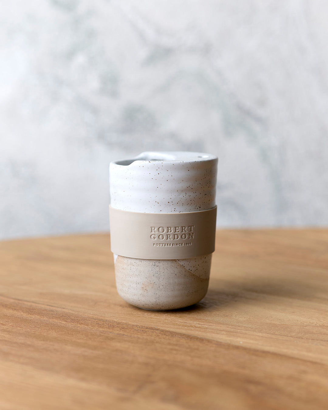 White speckle stoneware travel mug with lid and beige silicone band, on a timber bench