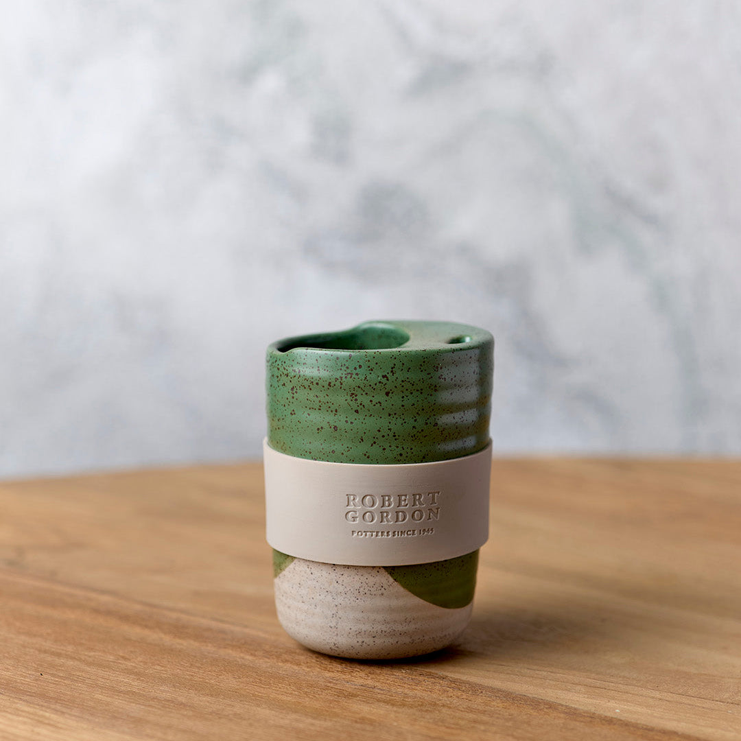 Green dip-glazed stoneware travel mug with beige silicone band, on a timber bench