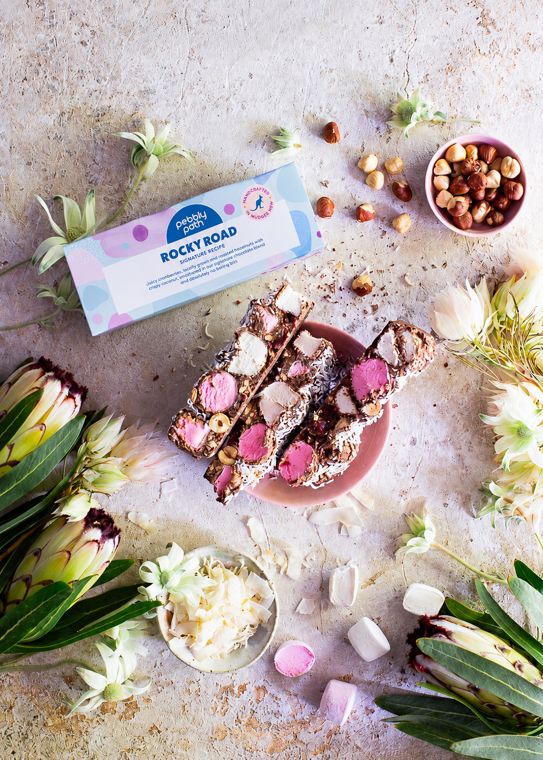 Pebbly Path Signature Recipe rocky road 100 g box with a cut slab on a pink plate, hazelnuts and coconut