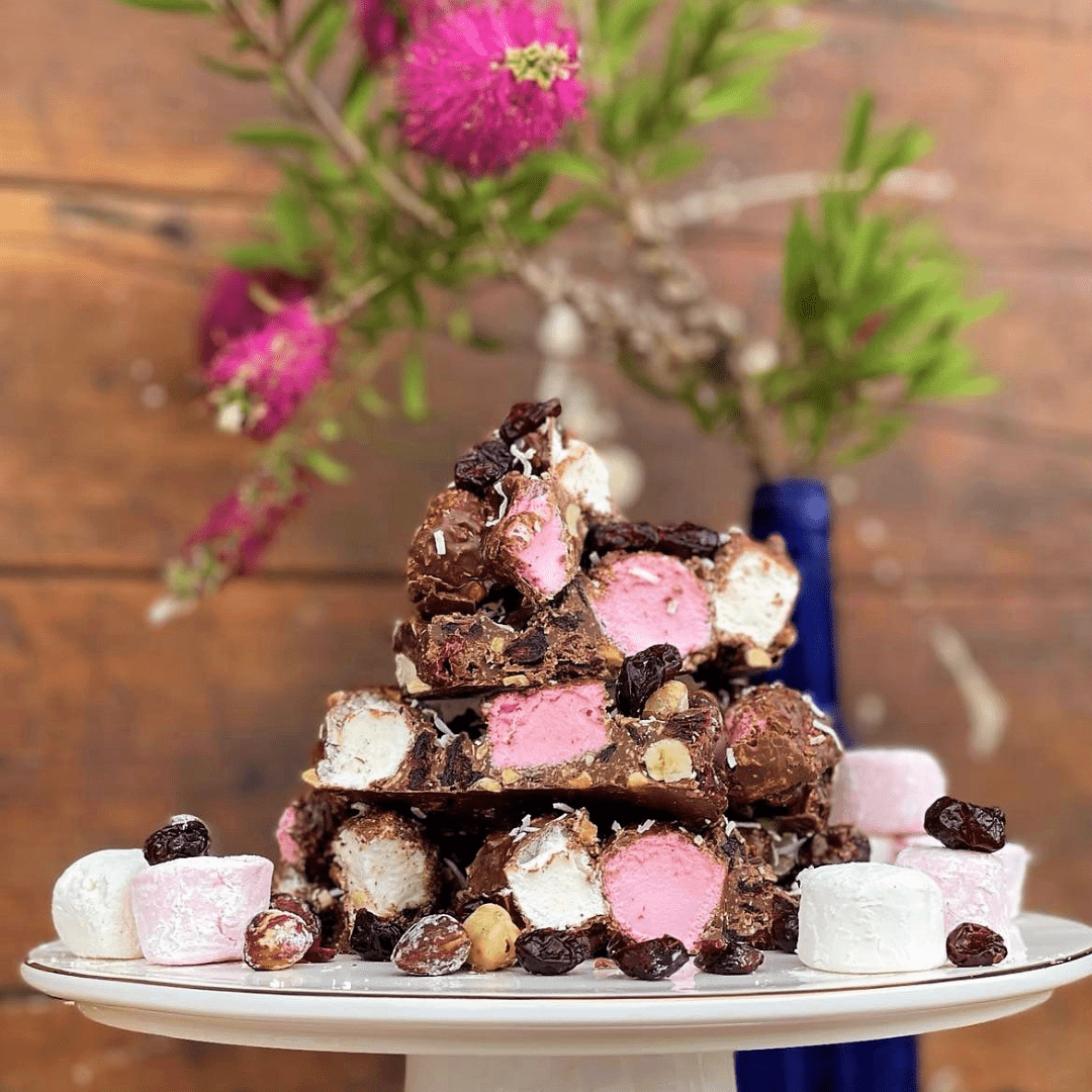 Signature Recipe rocky road stacked on a white cake stand with marshmallows and cranberries, pink bottlebrush flowers behind