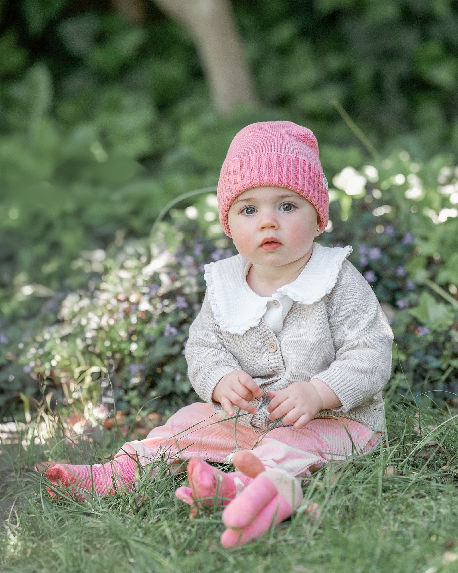 Baby sitting in a garden wearing the winter rose beanie with a cream knit cardigan
