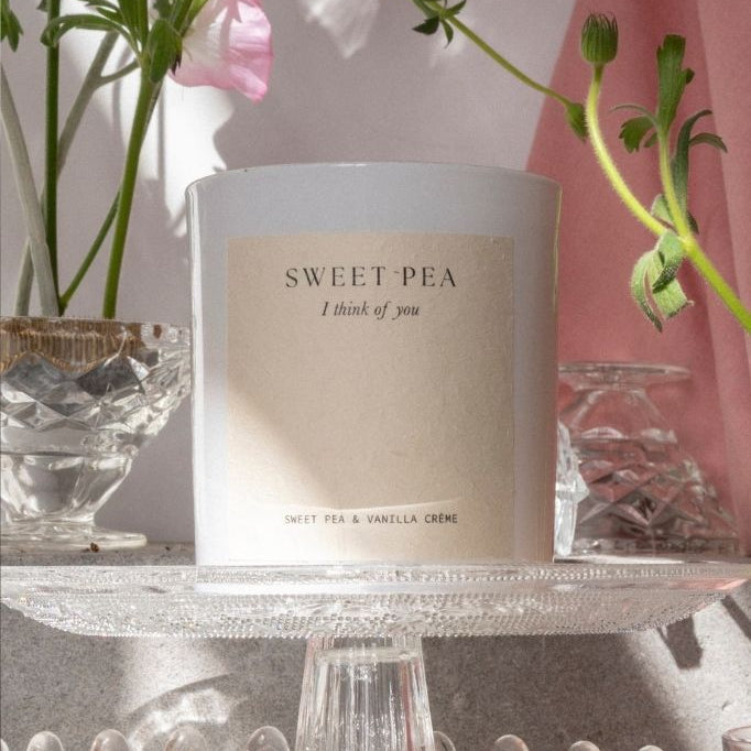 The Sweet Pea candle on a glass cake stand with sweet pea stems behind.