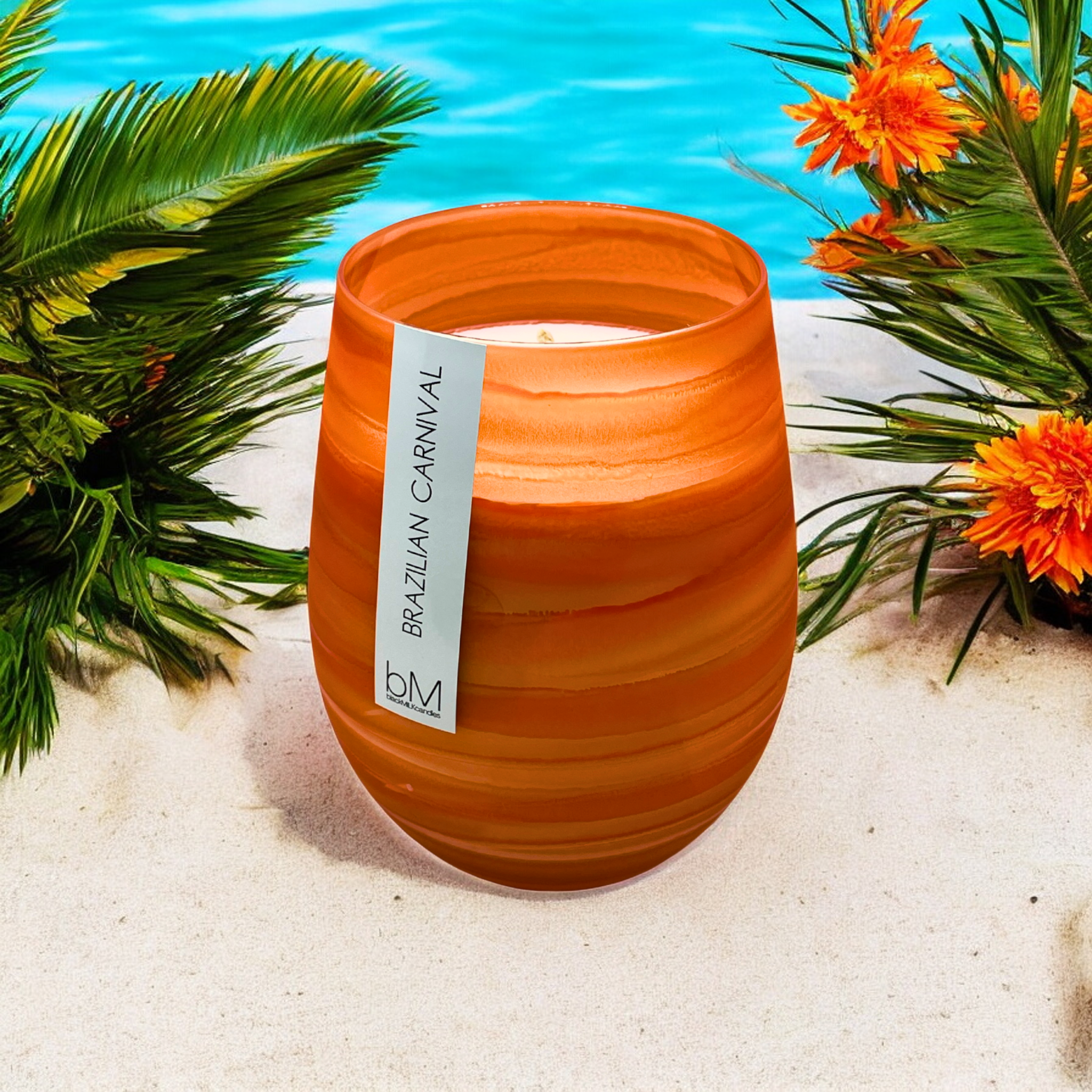 Wave Orange Brazilian Carnival candle on white sand by a pool, with palm fronds and orange blooms