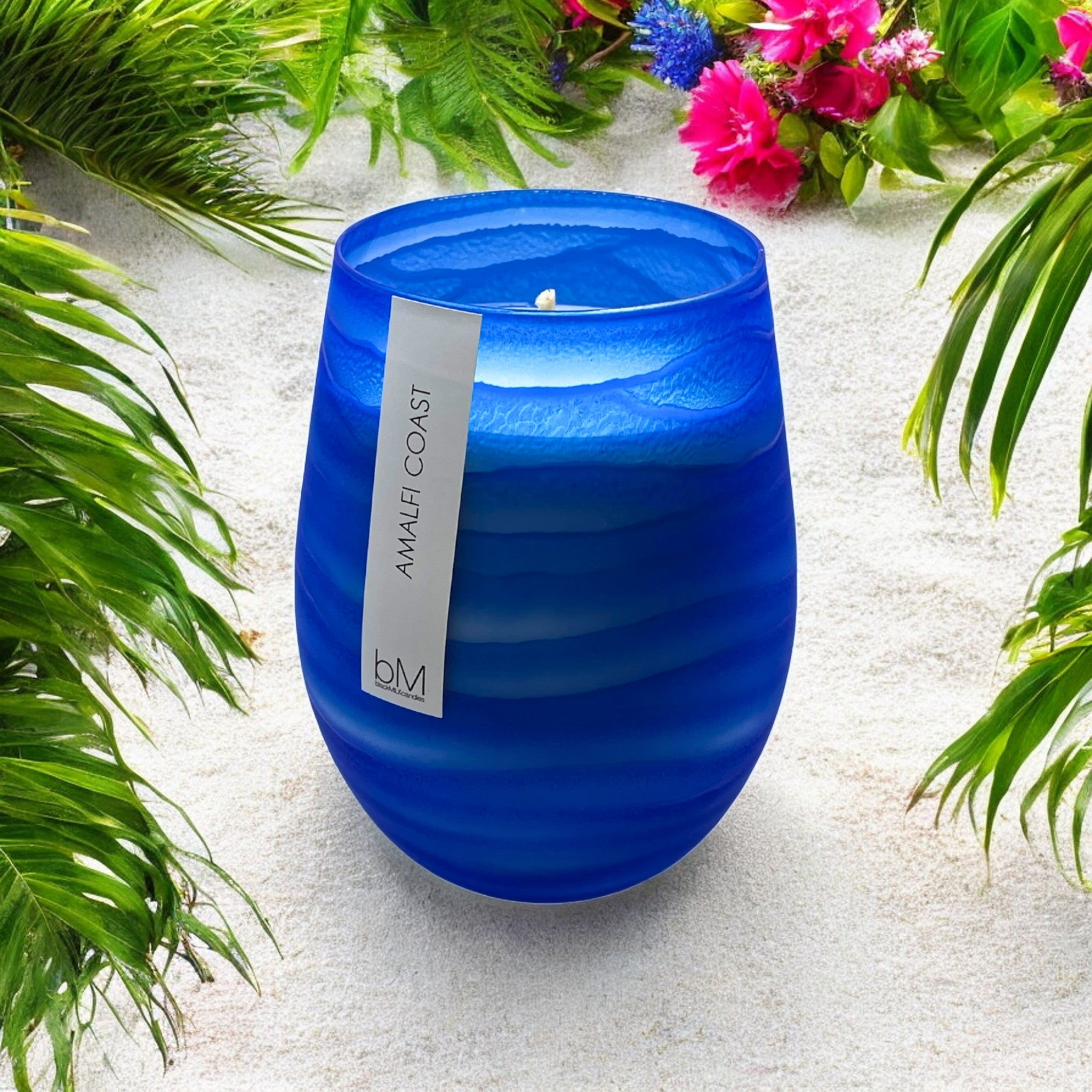 Wave Blue Amalfi Coast candle standing on white sand framed by palm fronds and pink tropical flowers