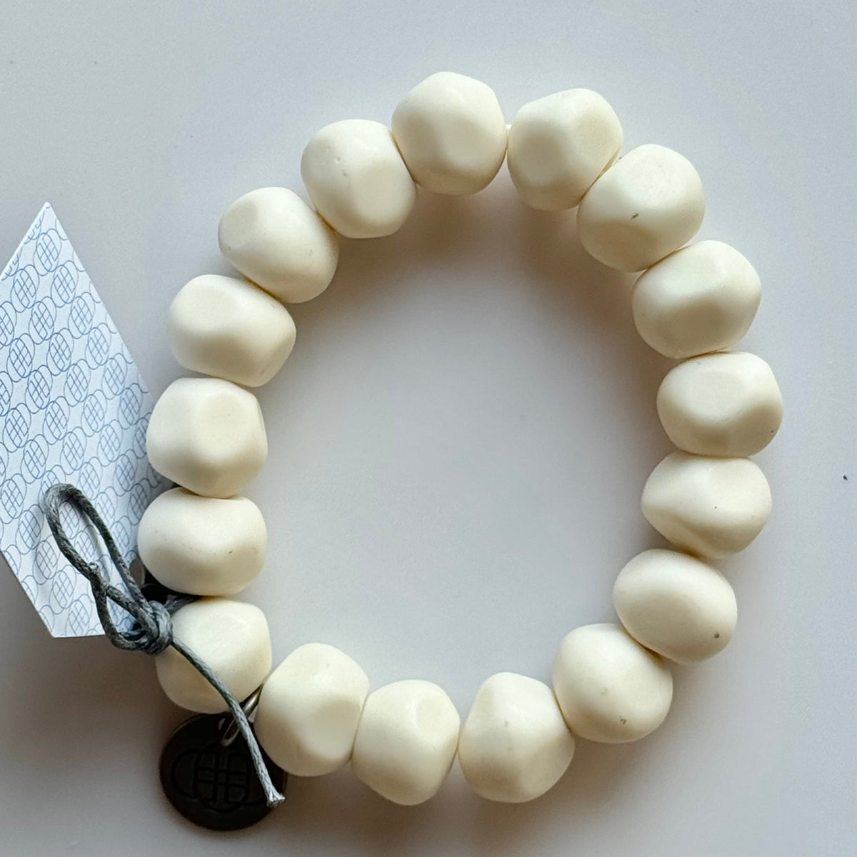 Rock bracelet in White: a stretch bracelet of chunky creamy white resin beads with its swing tag and metal charm, laid on a pale surface.