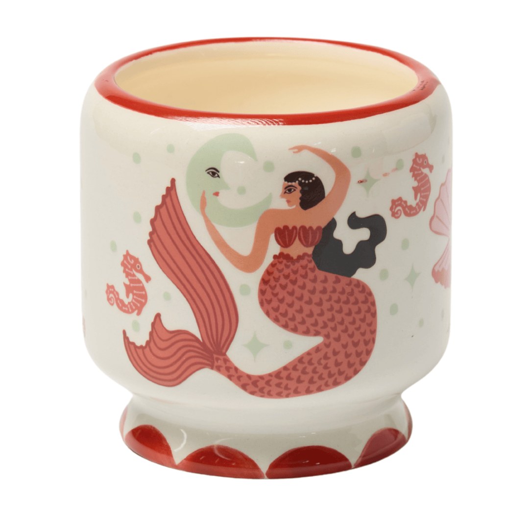 The Mermaid & Moon candle close up: a hand-painted coral mermaid on the cream ceramic pot, red at the rim and foot.
