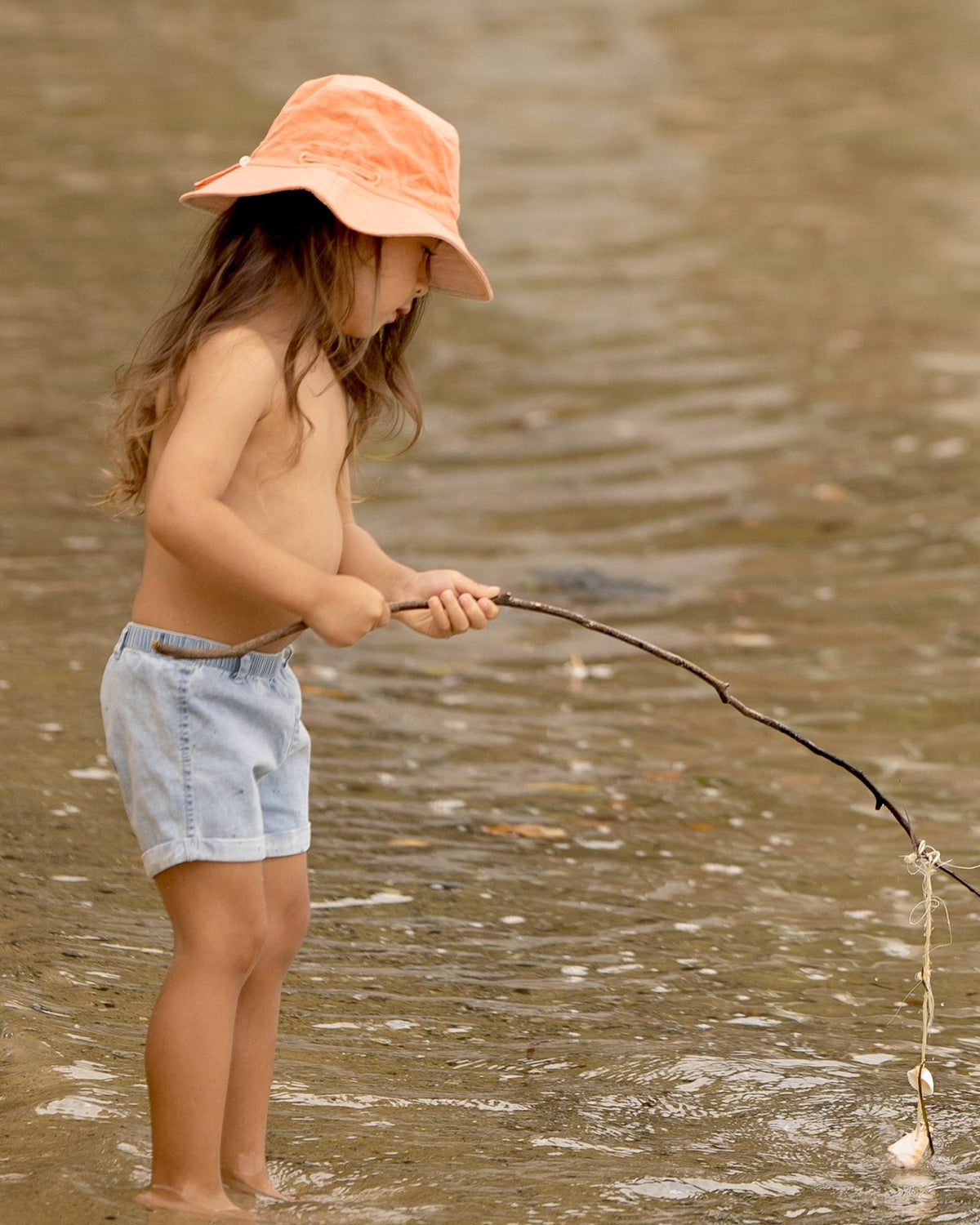 Child standing in shallow water with a stick, wearing the Indiana shorts and an orange sunhat
