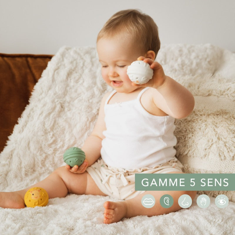 Baby sitting on a cream blanket holding the green ball, with the yellow ball beside their leg and a five-senses caption across the lower right.