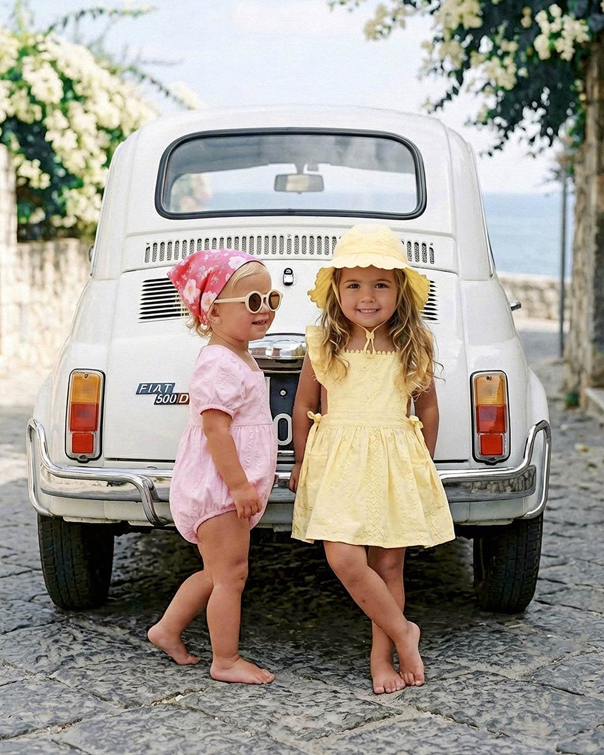 Toddler in the lemon Florence bell sunhat and matching pinafore beside a friend, in front of a white vintage car
