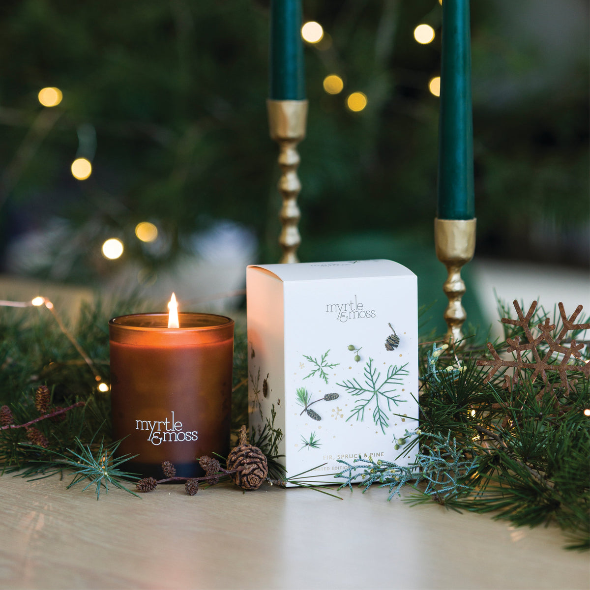 Lit Myrtle & Moss Fir, Spruce & Pine candle in an amber glass jar beside its white box printed with green pine snowflakes and cones, among pine branches.