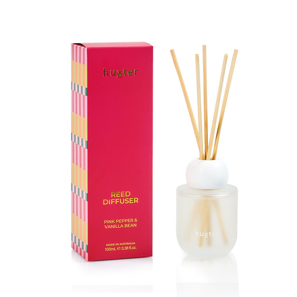 Red Huxter reed diffuser box with a striped pink, peach and green side band, beside a frosted glass bottle with a round white top and natural reeds.