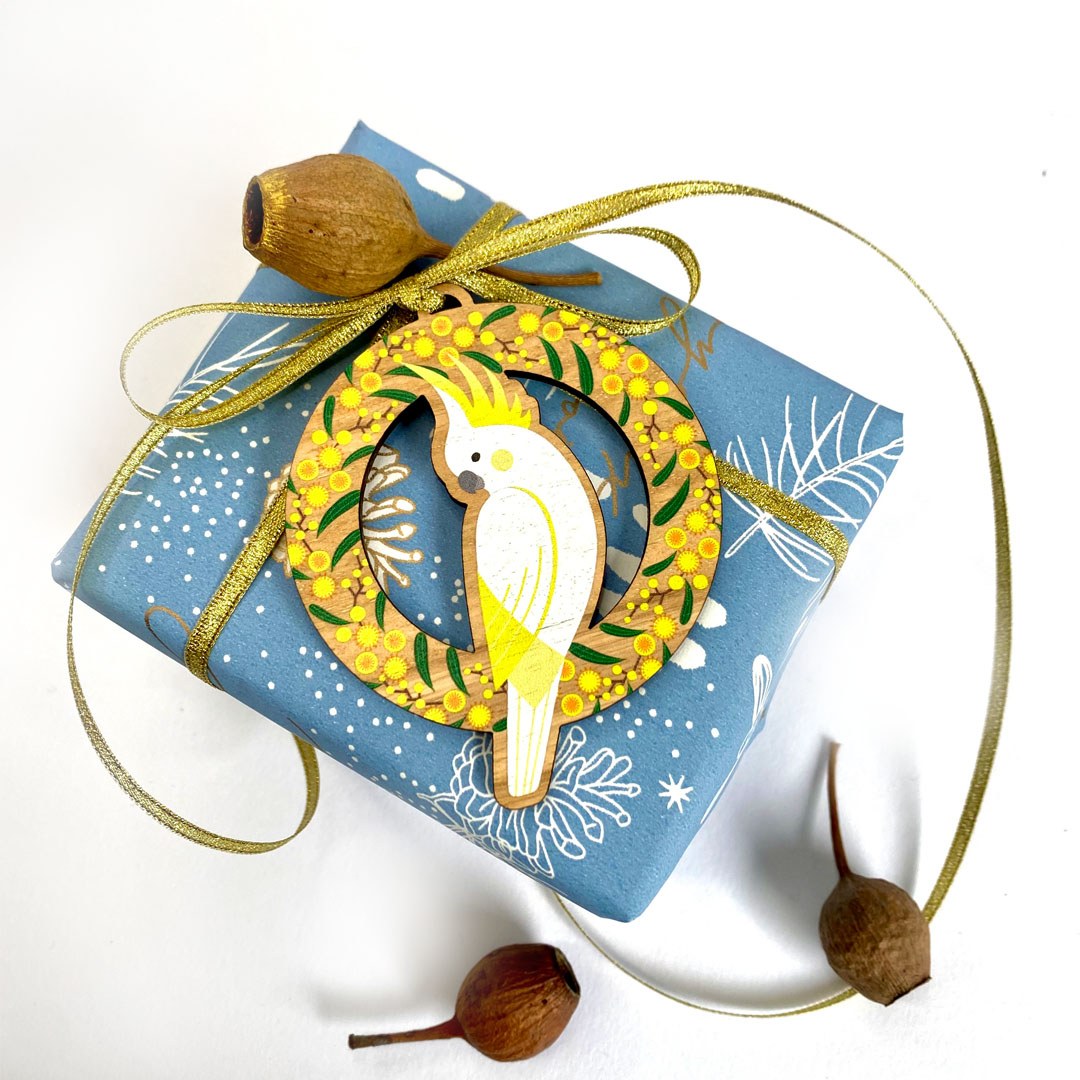 The Cockatoo ornament tied with gold ribbon onto a blue wrapped gift, gum nuts beside it.