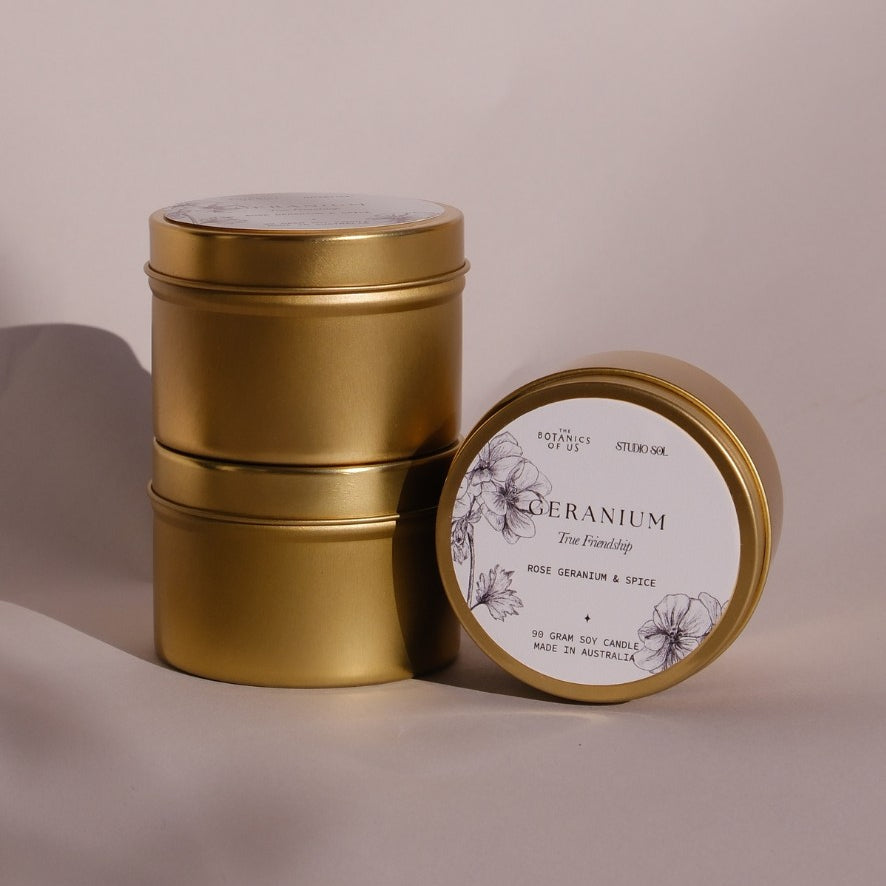 Three gold Geranium travel tins, one tipped forward to show its lid label with a drawing of the geranium.