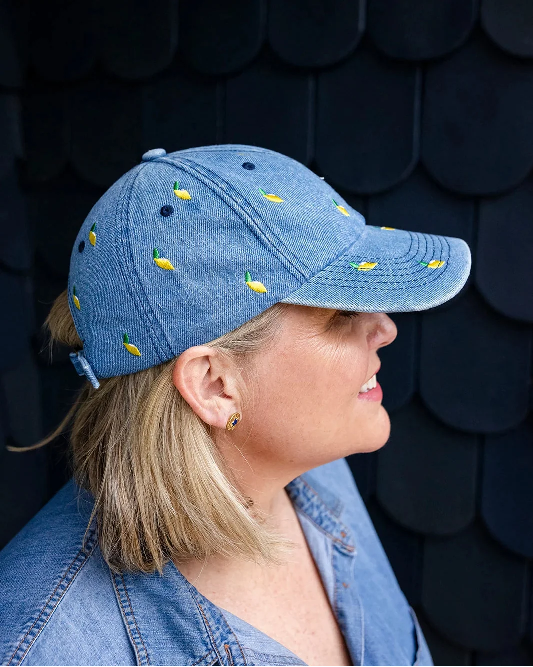 Woman in a denim shirt wearing the yellow-lemon denim cap in profile against a dark shingled wall.