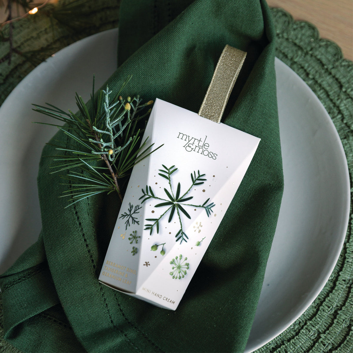Myrtle & Moss Bergamot hanging hand cream box with a gold ribbon loop, printed with green pine snowflakes and seed heads, on a green napkin.