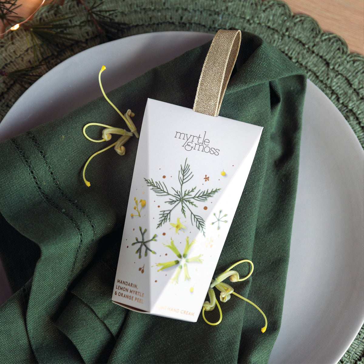 Myrtle & Moss Citrus hanging hand cream box with a gold ribbon loop, printed with green pine snowflakes and yellow flowers, on a green napkin.